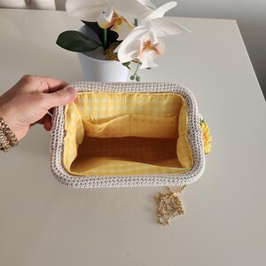 May include: Open, rectangular crochet handbag with a yellow gingham lining. The bag has a light gray exterior, a gold chain strap, and a yellow sunflower accent. The interior features two pockets.