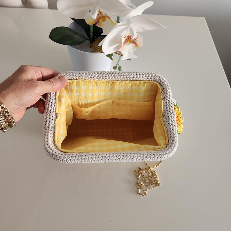 May include: Open, rectangular crochet handbag with a yellow gingham lining. The bag has a light gray exterior, a gold chain strap, and a yellow sunflower accent. The interior features two pockets.