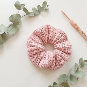 May include: A light pink crocheted hair scrunchie with a ribbed texture. The scrunchie is positioned next to a 4 mm crochet hook and sprigs of eucalyptus leaves. The crochet hook features a light-coloured handle with the text Tığ 4 mm.