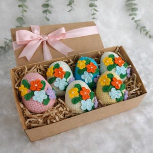 Pode incluir: Uma caixa de presente com seis ovos de Páscoa de crochê. Os ovos são decorados com flores coloridas em tons de laranja, amarelo e azul. A caixa é marrom com uma fita rosa.