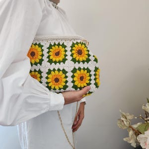 May include: A handmade crochet clutch featuring a sunflower design. The bag has a white base with yellow and brown sunflowers, green leaves, and a gold chain strap. The clutch is held by a person wearing a white blouse.