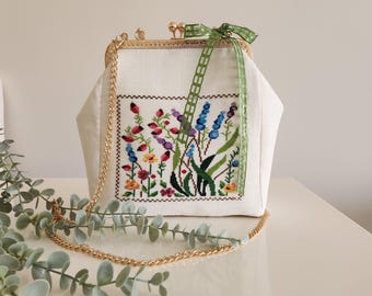 Könnte beinhalten: Eine weiße Stoff-Clutch mit einer goldenen Kettenkette und einem floralen Stickmuster. Die Stickerei zeigt eine Vielzahl von bunten Blumen, darunter blaue, lila, rosa, gelbe und grüne.