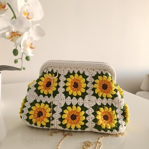 May include: A handmade crochet clutch featuring a sunflower design. The bag is cream-colored with yellow, brown, and green sunflower motifs. It has a gold chain strap and a clasp closure. The clutch is approximately 12 inches wide.