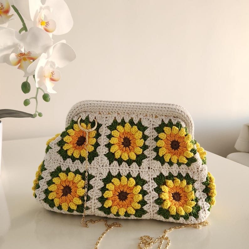 May include: A handmade crochet clutch featuring a sunflower design. The bag is cream-colored with yellow, brown, and green sunflower motifs. It has a gold chain strap and a clasp closure. The clutch is approximately 12 inches wide.