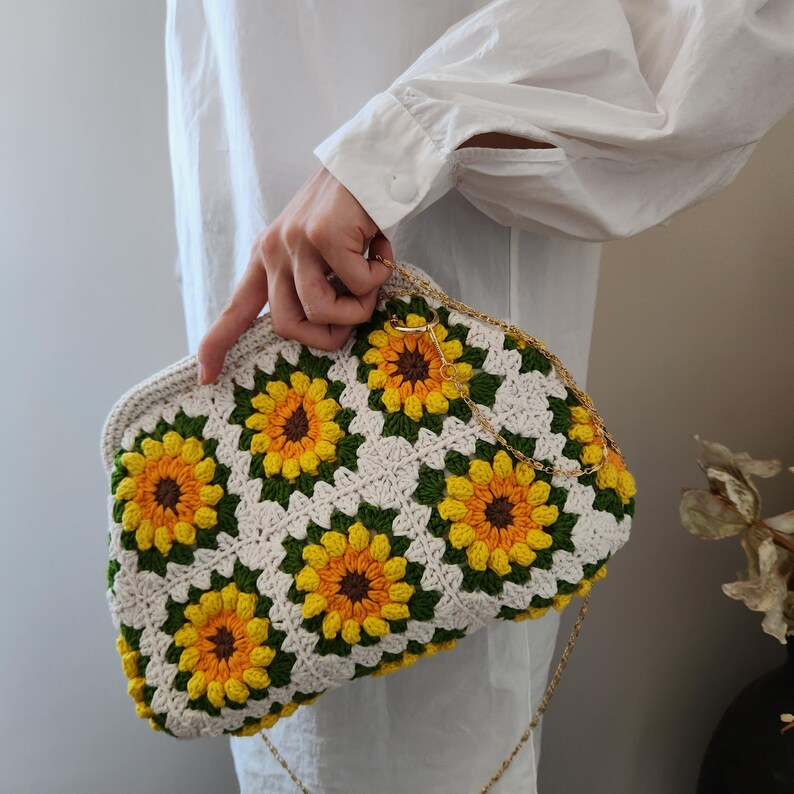 May include: A crochet clutch featuring a sunflower design. The bag is cream-colored with yellow and orange sunflowers, green leaves, and a gold chain strap. The clutch is held by a person wearing a white shirt.