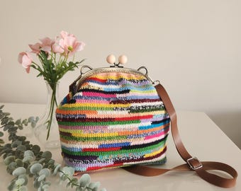 May include: A colorful, handmade crochet purse with a metal frame and wooden ball clasp. The bag features a brown leather strap and is filled with horizontal stripes of various colors. A vase of pink flowers sits in the background.