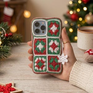 May include: A smartphone case covered in a crocheted granny square pattern featuring alternating red, green, and white yarn colors. The case includes a small, white, crocheted snowflake charm attached to the side, adding a festive detail.