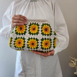 May include: A crochet clutch featuring a sunflower design. The bag is made with white, yellow, green, and brown yarn. It has a gold chain strap and a rectangular shape.