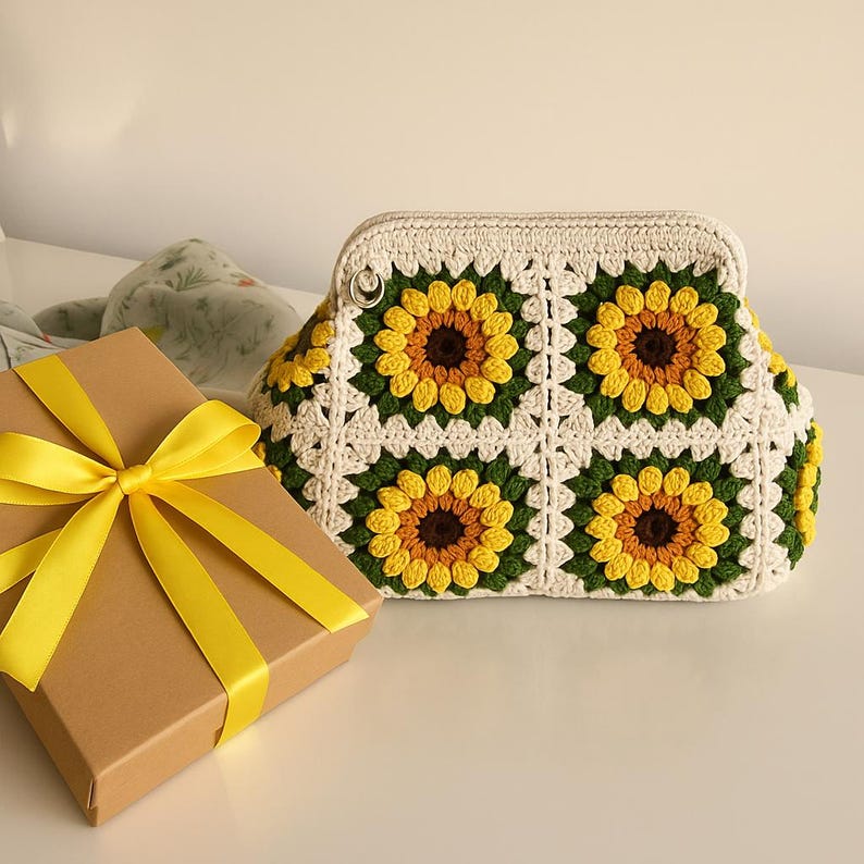 May include: A cream-colored crochet handbag with a sunflower pattern. The bag features yellow, brown, and green floral designs. A brown gift box with a yellow ribbon is in the foreground.