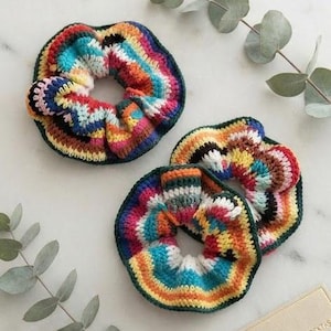 May include: Two circular hair scrunchies crafted from multicolored yarn in a crochet pattern. The scrunchies feature vibrant stripes of red, orange, yellow, green, blue, and white, with a dark green border along the outer and inner edges.