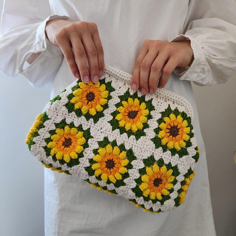 May include: A crocheted clutch featuring a sunflower design. The bag is white with yellow, green, and brown sunflower motifs. The clutch is held by a person wearing a white long-sleeved shirt.