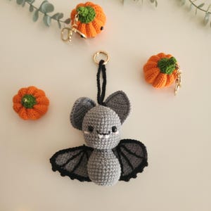 May include: A crocheted gray bat with black wings and a smiling face, hanging from a gold keyring. Two crocheted orange pumpkins with green stems and gold keyrings are also present, along with a third pumpkin.