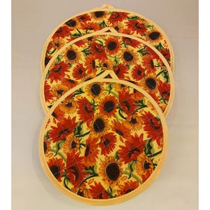 Op de afbeelding: Drie ronde, gestikte onderzetters met een zonnebloemmotief. De onderzetters zijn geel met rode en oranje zonnebloemen. De onderzetters zijn in elkaar genest.