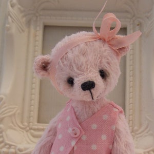 May include: A pink teddy bear wearing a pink dress with white polka dots and a pink bow. The bear is sitting on a white surface.