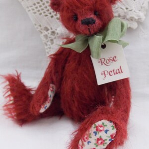 May include: A handmade red teddy bear with a green ribbon around its neck and a tag that reads "Rose Petal." The bear's paws and inner ears feature a floral fabric. The bear is a plush toy.