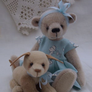 May include: A light brown teddy bear wearing a blue dress with white floral print and a blue bow. The bear is sitting with a light brown puppy with a blue bow.