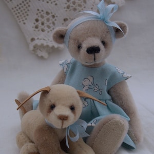 May include: A light blue and white dress with floral print is worn by a beige teddy bear with a blue bow. A beige puppy with a blue bow sits next to the bear. The image is captioned "Essential Bears".