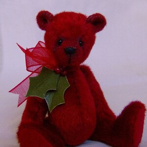 May include: A small, red teddy bear wearing a green holly leaf and a red ribbon. The bear is sitting on a white surface.