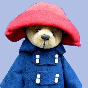 May include: A plush teddy bear wearing a blue coat with a hood and a red hat. The bear has brown fur and a black nose. The coat has four white buttons and two pockets. The text "Essential Bears" is visible at the bottom of the image.