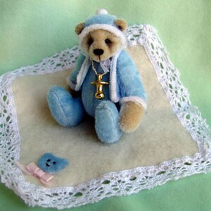 May include: A small blue teddy bear wearing a white hat and a gold necklace with a pacifier charm. The bear is lying on a white crocheted blanket.