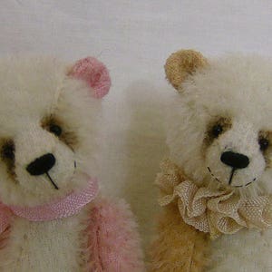 May include: Two white teddy bears with embroidered faces. One bear is wearing a pink ribbon and the other is wearing a beige ribbon.