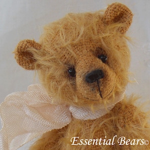 Può includere: Primo piano di un orsacchiotto marrone chiaro con una sciarpa bianca. L'orso ha un naso cucito e occhi neri. L'orso indossa una sciarpa bianca con una texture delicata. Il testo "Essential Bears" è visibile nell'angolo in basso a destra dell'immagine.