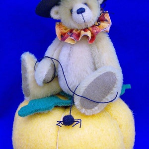 Puede incluir: Un oso de peluche con un sombrero de bruja negro y una bufanda colorida se sienta en una calabaza amarilla con una araña negra.