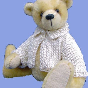 May include: A cream-colored teddy bear wearing a white knitted cardigan and a pink bow. The bear is sitting with its legs crossed and has a sweet expression on its face.