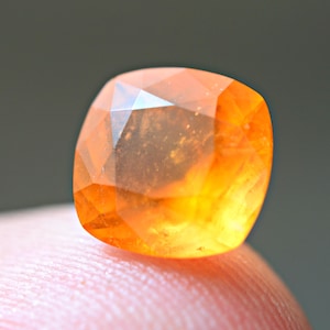 Puede incluir: Una gema naranja facetada con forma cuadrada. La gema tiene una superficie brillante y reflectante, que muestra una gama de tonos naranjas. La gema descansa sobre un dedo, con un fondo borroso.