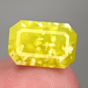 May include: A faceted, rectangular gemstone with a bright yellow hue and internal white inclusions. The gem has a defined rectangular shape with beveled edges, and is held by a finger in the foreground. The gem's color is consistent throughout.