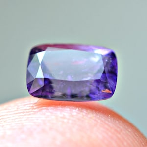 Puede incluir: Primer plano de una gema rectangular facetada de color púrpura intenso. La gema tiene una superficie pulida con múltiples facetas que reflejan la luz, creando un efecto brillante. La gema se sostiene en un dedo.
