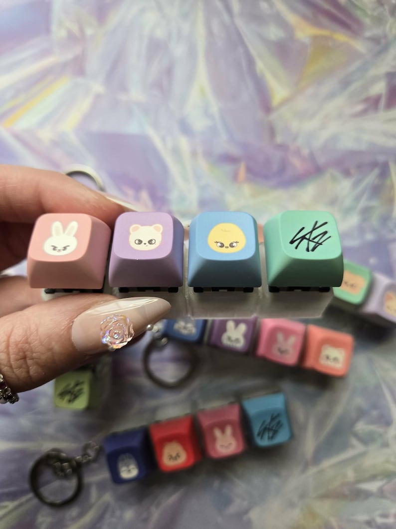May include: Pastel-coloured keyboard keycaps with animal and character designs. The keycaps are attached to keychains. Colours include pink, purple, blue, and green. The keycaps have a square shape.