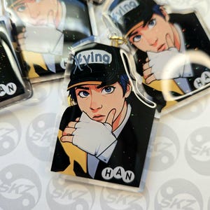 May include: Clear acrylic keychains featuring a stylized illustration of a person with blue eyes and blue hair wearing a black cap with the word "Xying". The keychains have a gold-colored chain and the name "HAN" is printed on the bottom.