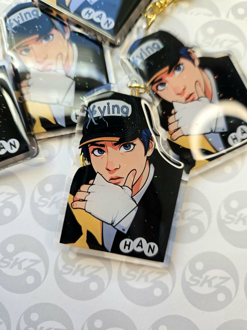 May include: Clear acrylic keychains featuring a stylized illustration of a person with blue eyes and blue hair wearing a black cap with the word "Xying". The keychains have a gold-colored chain and the name "HAN" is printed on the bottom.