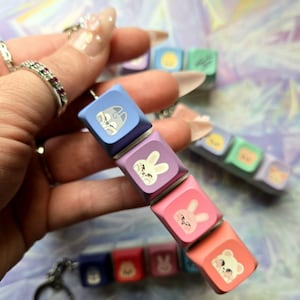 May include: A keychain with a series of colourful keyboard keycaps, each displaying a cartoon animal. The keycaps are in blue, purple, pink, and orange. The animal illustrations include a cat, rabbit, and bear.