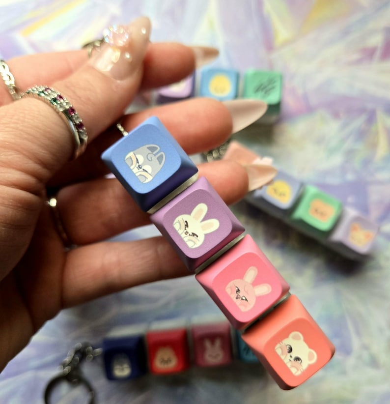 May include: A keychain with a series of colourful keyboard keycaps, each displaying a cartoon animal. The keycaps are in blue, purple, pink, and orange. The animal illustrations include a cat, rabbit, and bear.