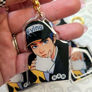May include: A clear acrylic keychain featuring a stylized anime-style illustration of a person wearing a black cap with the word "Xying" and a black suit. The keychain has a gold-colored chain and clasp. The name "HAN" is printed on the bottom.