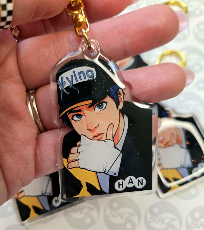 May include: A clear acrylic keychain featuring a stylized anime-style illustration of a person wearing a black cap with the word "Xying" and a black suit. The keychain has a gold-colored chain and clasp. The name "HAN" is printed on the bottom.