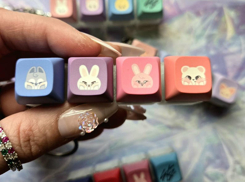 May include: Close-up of four colourful keyboard keycaps. The keycaps are square and feature cartoon animal designs: a rabbit, a squirrel, and a bear. The keycaps are in shades of blue, purple, and pink.