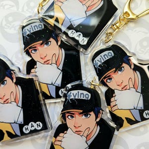 May include: Multiple clear acrylic keychains featuring a stylized illustration of a person wearing a black cap with the word "Xyving" and the name "HAN". The character is wearing a black suit and white gloves. Each keychain has a gold-colored keyring.