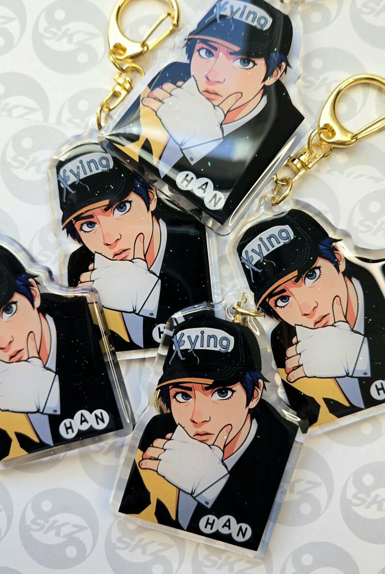 May include: Multiple clear acrylic keychains featuring a stylized illustration of a person wearing a black cap with the word "Xyving" and the name "HAN". The character is wearing a black suit and white gloves. Each keychain has a gold-colored keyring.