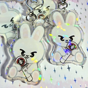 May include: Clear acrylic keychains featuring a white cartoon bunny holding a red and black object. The bunny has black eyes and eyebrows. The keychains have a silver-colored metal clasp and a holographic effect.