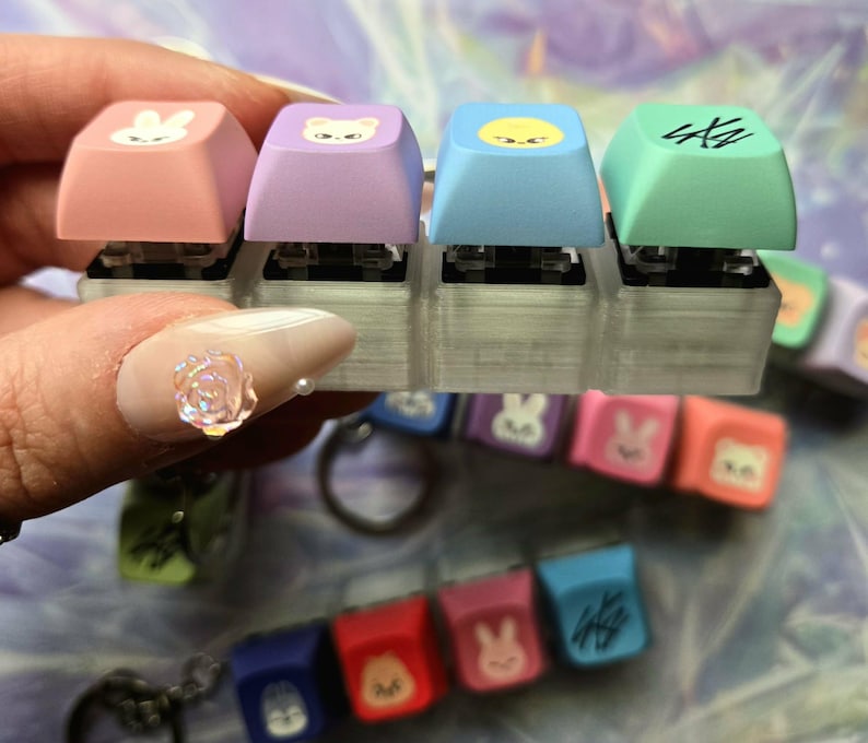 May include: A close-up of four brightly coloured keyboard keycaps. The keycaps are pink, purple, blue, and green, each with a different cartoon character design. Other keycaps and a keychain are visible in the background.