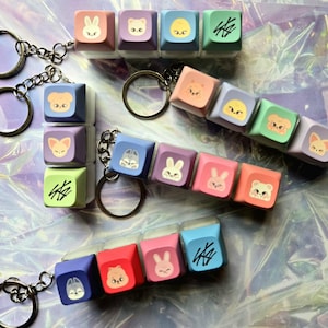 May include: Keyrings with colourful square charms, each displaying animal faces and symbols. The charms, in pastel shades like pink, purple, blue, and green, are attached to silver keyrings. The animal designs include a bunny, bear, and fox.