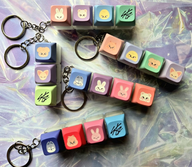 May include: Keyrings with colourful square charms, each displaying animal faces and symbols. The charms, in pastel shades like pink, purple, blue, and green, are attached to silver keyrings. The animal designs include a bunny, bear, and fox.
