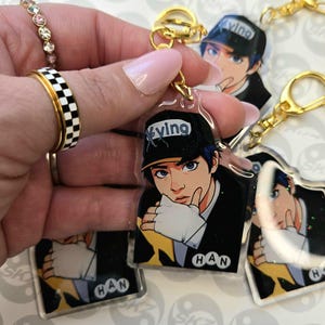 May include: Clear acrylic keychain featuring a cartoon illustration of a person with blue hair and a black cap with the word "Xying". The keychain has a gold-colored clasp and a small tag that says "HAN".