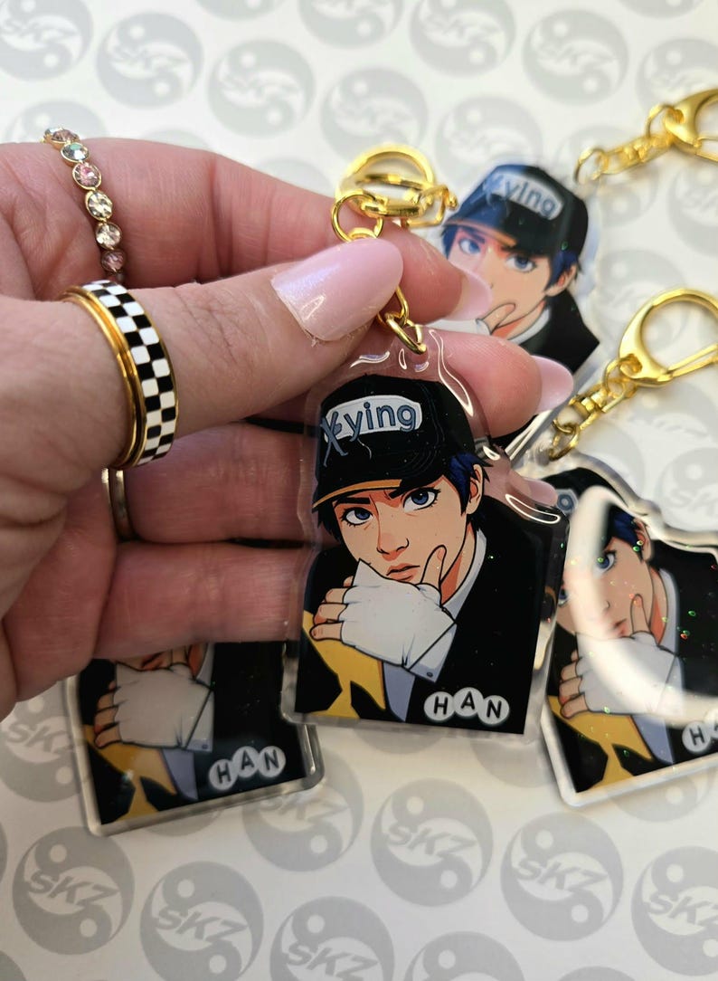 May include: Clear acrylic keychain featuring a cartoon illustration of a person with blue hair and a black cap with the word "Xying". The keychain has a gold-colored clasp and a small tag that says "HAN".