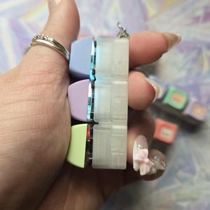 May include: A keyboard keycap pendant with pastel-coloured keycaps in light blue, lavender, and yellow-green. The pendant is held in a hand with a silver ring. The background is a blurred mix of purple and white.
