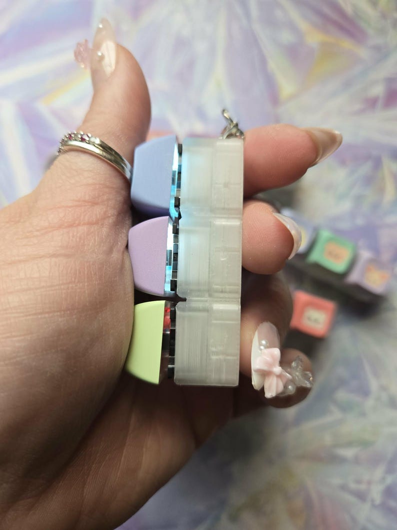 May include: A keyboard keycap pendant with pastel-coloured keycaps in light blue, lavender, and yellow-green. The pendant is held in a hand with a silver ring. The background is a blurred mix of purple and white.