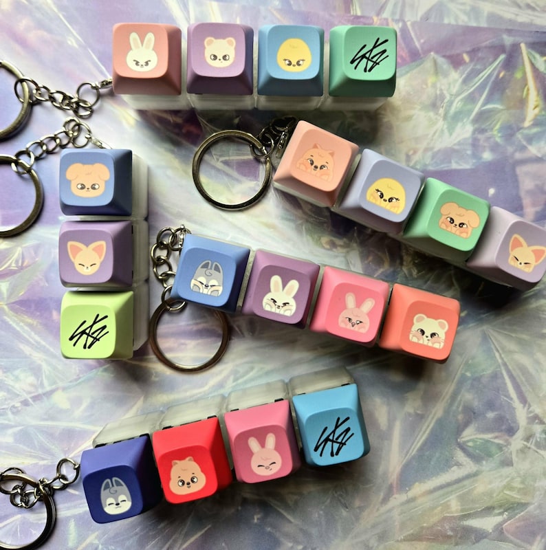 May include: A selection of colourful keycaps featuring cartoon animal faces and a stylized signature. The keycaps are attached to keychains. The keycaps are in shades of pink, blue, green, and purple.